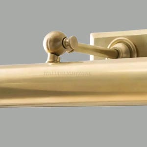 May include: Close-up of a brass-colored wall-mounted light fixture. The fixture has a rectangular base and a curved arm with a small, round adjustment knob. The text "ITALIANLIGHTZONE" is visible.