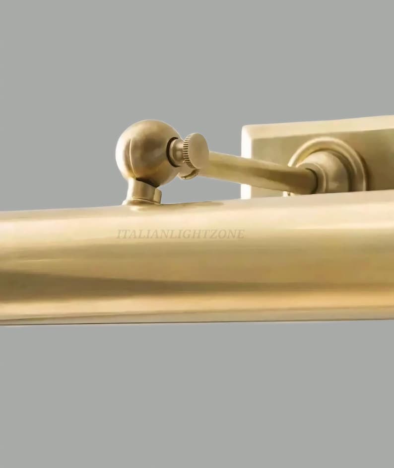 May include: Close-up of a brass-colored wall-mounted light fixture. The fixture has a rectangular base and a curved arm with a small, round adjustment knob. The text "ITALIANLIGHTZONE" is visible.
