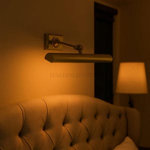 May include: A wall-mounted, brass-toned picture light illuminates a tufted headboard. The light has a rectangular base and a pivoting arm. The text "ITALIANLIGHTZ" is visible on the light fixture. A bedside lamp is in the background.