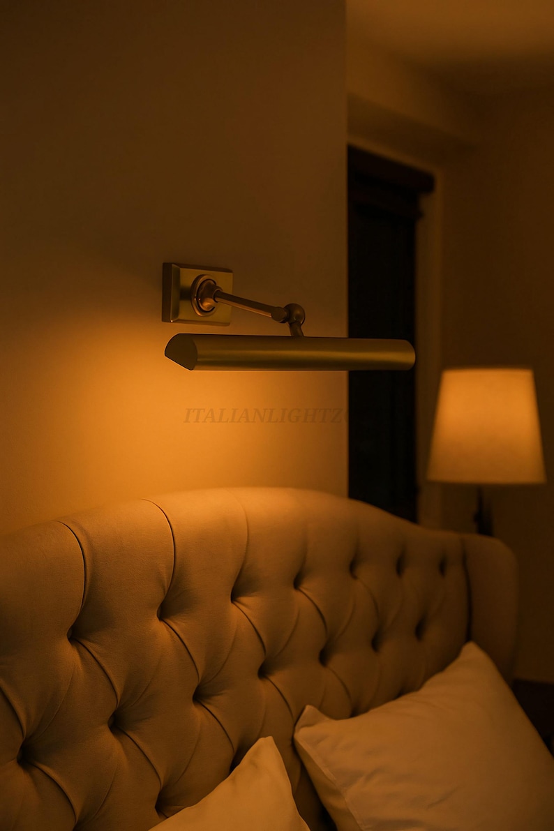 May include: A wall-mounted, brass-toned picture light illuminates a tufted headboard. The light has a rectangular base and a pivoting arm. The text "ITALIANLIGHTZ" is visible on the light fixture. A bedside lamp is in the background.