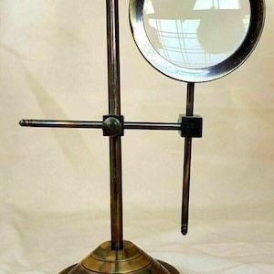 Op de afbeelding: Antieke messing vergrootglas op een standaard. De ronde lens wordt vastgehouden door een donker metalen frame. De standaard heeft een cirkelvormige basis en een verticale paal met verstelbare horizontale arm. Het vergrootglas is een vintage stijl.