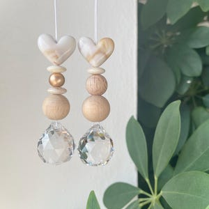 Könnte beinhalten: Zwei hängende Ornamente mit einer weißen herzförmigen Perle an der Spitze, gefolgt von einer kleinen cremefarbenen Scheibe, einer runden Perle, einer größeren Holzkugel und einer klaren, facettierten Kristallkugel an einer dünnen weißen Schnur.