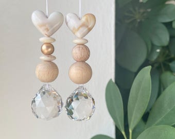 Puede incluir: Dos adornos colgantes con cuentas blancas en forma de corazón con patrones jaspeados, pequeñas cuentas de madera redondas y una cuenta dorada, terminados con esferas de cristal transparente facetado en hilos blancos.