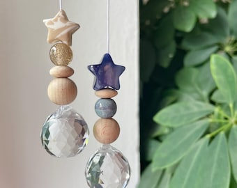 Puede incluir: Dos adornos colgantes en hilos blancos. El de la izquierda tiene una estrella veteada beige, una cuenta de purpurina dorada y dos cuentas de madera sobre una esfera de cristal facetado. El de la derecha tiene una estrella morada y cuentas.