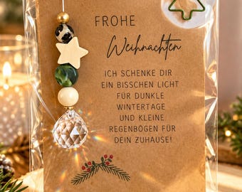 May include: A clear plastic-wrapped gift card on brown paper featuring the text "FROHE Weihnachten ICH SCHENKE DIR EIN BISSCHEN LICHT FÜR DUNKLE WINTERTAGE UND KLEINE REGENBÖGEN FÜR DEIN ZUHAUSE!" with a green tree-shaped paperclip and a hanging crystal bead ornament.