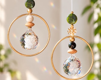 Puede incluir: Dos atrapasoles colgantes con anillos de metal dorado, colgantes de cristal transparente facetado y una disposición vertical de cuentas en tonos verde, blanco, marrón y negro, suspendidos por hilos blancos finos con reflejos.