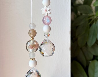 Puede incluir: Dos adornos colgantes con prismas de cristal transparente facetado en la base. Los hilos están compuestos por varias cuentas, incluyendo esferas blancas mate, una cuenta con forma de flor rosa, una esfera dorada brillante y detalles de vidrio rosa iridiscente.