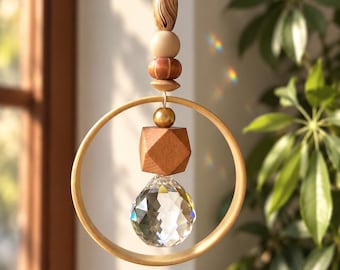 Puede incluir: Un atrapasol colgante con un aro de madera que rodea una esfera de cristal transparente facetada. Sobre el aro, una disposición vertical de cuentas incluye un óvalo jaspeado, una esfera lisa, una cuenta estriada y una esfera de tono dorado.