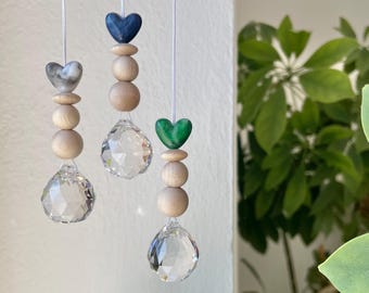 Puede incluir: Tres atrapasoles de cristal colgantes con cuentas de madera y detalles en forma de corazón en azul, verde y gris veteado. Los atrapasoles están suspendidos de cuerdas blancas, con un fondo de pared blanca y plantas verdes.