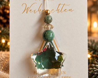 Puede incluir: Un adorno de cristal transparente en forma de estrella colgado de un cordón blanco, decorado con una pequeña cuenta dorada, una cuenta verde estampada, una cuenta dorada estriada y una cuenta verde más grande sobre una tarjeta blanca con texto dorado.