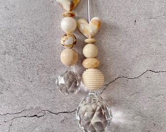 Puede incluir: Dos adornos colgantes con cuerdas blancas, cada uno decorado con una serie de cuentas. Las cuentas incluyen esferas de madera lisa, cuentas en forma de corazón jaspeadas en beige y blanco, y colgantes de cristal facetado en la base.