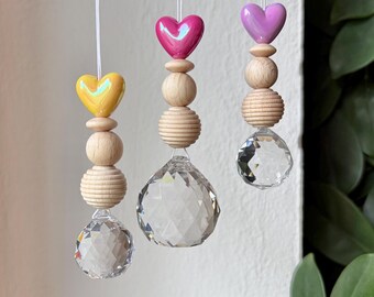Puede incluir: Tres adornos colgantes con esferas de cristal facetado transparente en la parte inferior, rematados con cuentas de madera apiladas en texturas lisas y estriadas, y finalizados con una cuenta brillante en forma de corazón amarilla, rosa y morada.