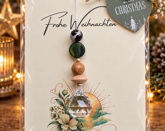 Puede incluir: Un adorno colgante decorativo con un cristal transparente facetado, dos cuentas de madera, una cuenta verde y una cuenta con patrón en blanco y negro. El adorno está sujeto a una tarjeta con el texto "Frohe Weihnachten" y una etiqueta de corazón "Merry Christmas".