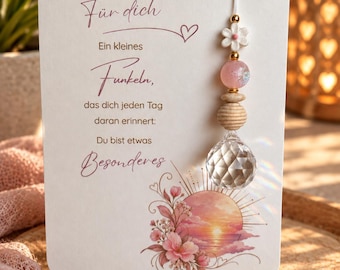 以下が含まれることがあります： ドイツ語のテキスト「Für dich Ein kleines Funkeln, das dich jeden Tag daran erinnert: Du bist etwas Besonderes」と夕日のイラストが描かれた白い長方形のカード。透明なクリスタルプリズムが付いたビーズのオーナメントがカードから吊り下げられています。