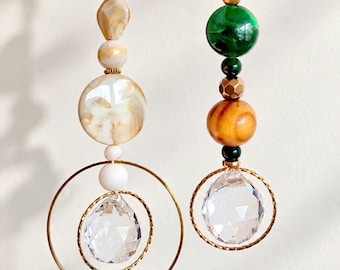 Puede incluir: Dos adornos colgantes con cuentas en tonos crema, beige, verde y marrón. Cada adorno incluye un anillo de metal dorado y un colgante de cristal transparente facetado en forma de lágrima suspendido de un cordón blanco.