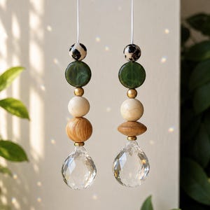 Könnte beinhalten: Zwei vertikale Hängedekorationen mit einer Perlenschnur in verschiedenen Texturen und Farben, darunter gesprenkeltes Weiß, Dunkelgrün, Metallic-Gold, Creme und Holzmaserung, die in einem klaren, facettierten, tropfenförmigen Glaskristall enden.