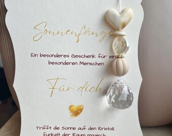 Puede incluir: Una tarjeta blanca con bordes festoneados y texto dorado que dice Sonnenfänger, Ein besonderes Geschenk für einen besonderen Menschen, Für dich, y Trifft die Sonne auf den Kristall, funkelt der Raum magisch. Lass dich verzaubern! Un atrapasoles de cristal cuelga de la tarjeta.