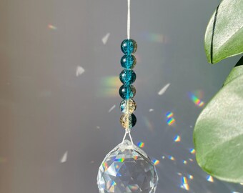 Puede incluir: Un atrapasoles colgante con una esfera de cristal transparente y facetada suspendida por un hilo blanco. Sobre la esfera, una fila vertical de cinco cuentas redondas presenta un degradado de color desde azul cerceta a dorado.