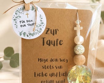 May include: A baptism gift set in a clear plastic sleeve featuring a brown card with the text Zur Taufe and a German blessing. Attached is a round tag with eucalyptus leaves and a cross, plus a hanging ornament with beads and a crystal prism.