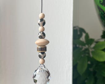 Puede incluir: Un adorno colgante con una gota de cristal transparente y facetada suspendida de un cordón negro. El diseño incluye una disposición vertical de pequeñas cuentas de madera redondas, dos cuentas redondas con un patrón de manchas blancas y negras, una cuenta de madera plana y una cuenta cilíndrica marrón.