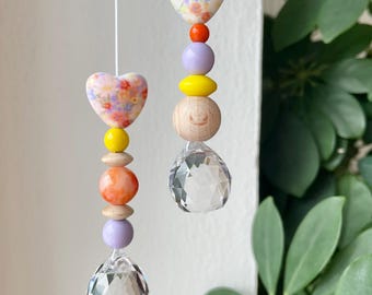 Puede incluir: Dos adornos colgantes con cuerdas blancas, cada uno decorado con una cuenta en forma de corazón con estampado floral, cuentas de madera redondas en amarillo, naranja y lavanda, y un prisma de cristal transparente facetado en forma de gota.