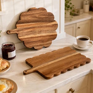 May include: Two wooden serving boards with scalloped edges and handles. One board is round and stands upright, while the other is rectangular and lies flat on the counter. Both boards feature a natural wood grain pattern in medium brown tones.
