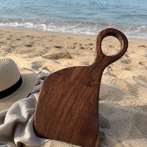 May include: A dark brown wooden cutting board with a curved edge and a circular handle cutout, positioned next to a beige straw hat with a black band, resting on a sandy surface with the ocean visible in the background.