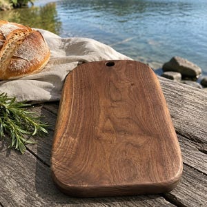 May include: A rectangular wooden cutting board with rounded corners and a small circular hole at the top center. The board features a rich, dark brown wood grain pattern and a smooth, polished surface, presented as a functional kitchen tool.