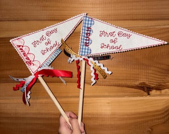 May include: Two triangular fabric pennants on wooden sticks, each embroidered with the text First Day of School in red. One pennant features the name Ramsey in red, while the other features the word RUSH in blue. Both are decorated with ribbons.
