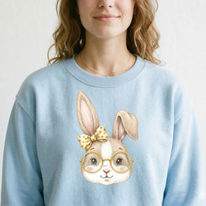 Puede incluir: Sudadera azul claro con la cara de un conejo de dibujos animados. El conejo tiene gafas y un lazo amarillo con lunares. La sudadera es de color azul claro liso.