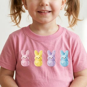 Puede incluir: Camiseta rosa con cuatro gráficos de conejitos coloridos en rosa, amarillo, morado y azul. Los conejitos tienen pequeños ojos negros y están dispuestos horizontalmente en la parte delantera. Cuello redondo.
