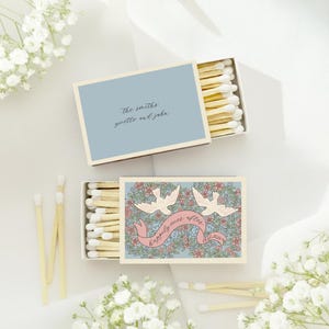 Puede incluir: Dos cajas de cerillas abiertas con dise&ntilde;os florales y de palomas. Una caja tiene el texto "The Smiths Yoette and John" sobre un fondo azul. La otra caja tiene el texto "happily ever after" en una pancarta rosa con dos palomas.