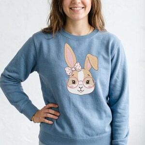 Puede incluir: Sudadera azul claro con un diseño bordado de cara de conejo. El conejo tiene orejas marrones con interiores rosas, un lazo rosa y blanco y gafas redondas. La sudadera es de color azul claro liso.