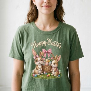 May include: Green t-shirt with a watercolor illustration featuring two bunnies, a basket of eggs, and the words "Happy Easter." The bunnies are brown with pink accents, and the basket is adorned with a pink bow. Colorful Easter eggs are scattered around the bunnies.