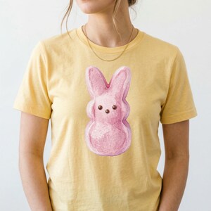 Puede incluir: Camiseta amarilla con una ilustración de un conejito de malvavisco rosa. El conejito tiene ojos marrones y está centrado en la camiseta. La camiseta tiene mangas cortas y cuello redondo.