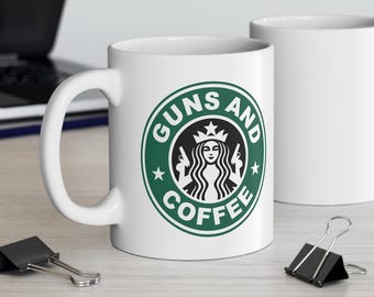 May include: Two white ceramic coffee mugs. One mug features a green and white logo that reads "Guns and Coffee" with a woman holding guns. The other mug is plain white. The mugs are on a light-colored surface.
