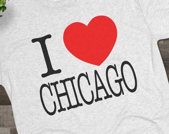 May include: Light gray t-shirt with the phrase "I ❤️ CHICAGO" printed in black and red. The shirt is displayed with black canvas shoes and eyeglasses on a wooden surface. The shirt is a casual wear item.