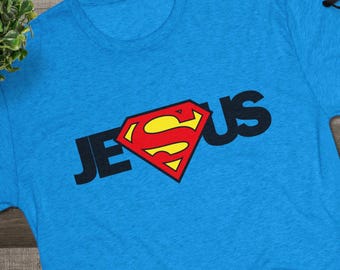 May include: A bright blue short-sleeved t-shirt featuring the black text JESUS printed across the chest. The letter U is replaced by a red and yellow diamond-shaped emblem with a stylized S in the center. The shirt has a crew neckline.