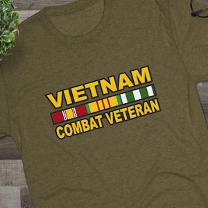 Pode incluir: Camiseta verde-oliva com o texto "VIETNAM COMBAT VETERAN" em amarelo e branco. A camiseta apresenta um design de fitas coloridas. Tênis preto e branco e óculos também são visíveis.