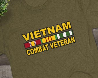May include: Olive green t-shirt with the text "VIETNAM COMBAT VETERAN" in yellow and white. The shirt features a design of colored ribbons. Black and white sneakers and eyeglasses are also visible.