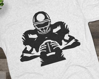 May include: Light gray t-shirt featuring a black graphic of a football player in a helmet, holding a football. The shirt is made of a soft material and is suitable for casual wear. The design is a bold, simple illustration.