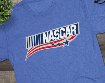 May include: A blue t-shirt with the word "NASCAR" in white, outlined in navy blue, with a red and white striped design and three blue stars. The shirt is paired with black and white sneakers and glasses.
