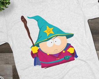 May include: A light gray heathered t-shirt featuring a graphic of a cartoon character wearing a teal wizard hat with a yellow star, a purple robe, and holding a brown wooden staff. The character has a round face and is depicted in a flat style.