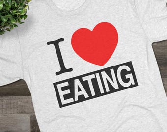 May include: A light gray heathered short-sleeve t-shirt featuring the black text I and the word EATING inside a black rectangular box, separated by a large red heart symbol. The shirt is displayed with black sneakers and dark-rimmed glasses.
