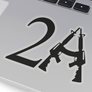 以下が含まれることがあります： 黒色のステッカーで、数字の「2A」と2丁のライフルのシルエットが描かれています。ステッカーは銀色のノートパソコンに貼られており、キーボードが見えます。大胆なグラフィックデザインです。