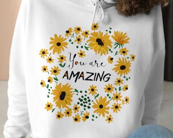 May include: White cropped hoodie with a sunflower design and the text "You are AMAZING". The design features yellow sunflowers and green leaves arranged in a circular pattern. The hoodie has a drawstring and is worn with blue jeans.