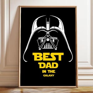 May include: Framed art print featuring a stylized white illustration of Darth Vader's helmet on a black background. The words "BEST DAD IN THE GALAXY" are in yellow. The artwork is in a wooden frame.