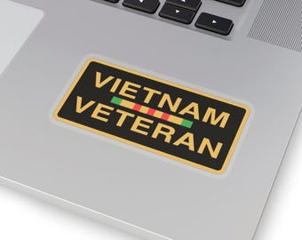 May include: A rectangular sticker with the words "VIETNAM VETERAN" in yellow text on a black background. The sticker has a gold border and a horizontal stripe of green, red, and yellow. The sticker is placed on a silver laptop.