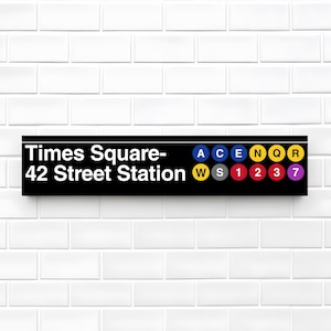 以下が含まれることがあります： 「Times Square - 42 Street Station」と白字で書かれた黒い長方形の看板。A、C、E、N、Q、R、W、S、1、2、3、7などの文字と数字が書かれた色の付いた円も含まれています。背景は白いレンガの壁です。