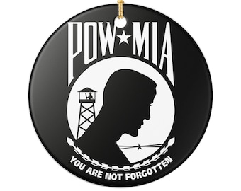 May include: A round black ceramic ornament featuring the white POW-MIA logo. The design includes a silhouette of a person, a guard tower, and the text POW-MIA with a star in the center. Below, the text reads YOU ARE NOT FORGOTTEN.
